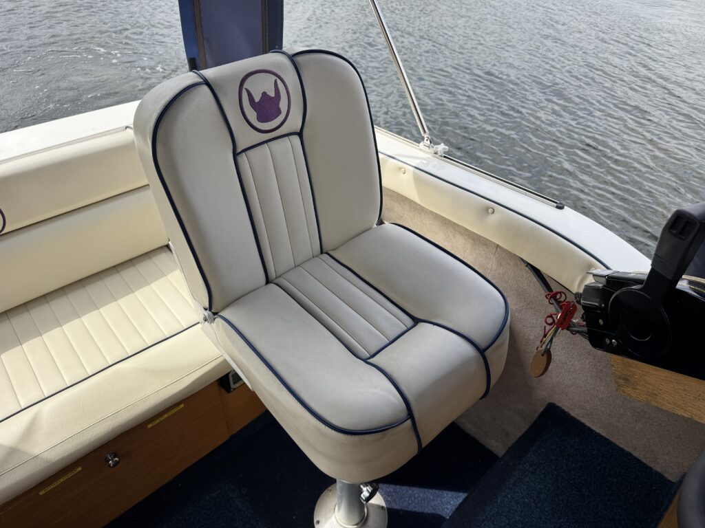 A white and gray boat seat with vertical stitching and a purple emblem on the back. The seat is positioned on a boat, with a portion of the boat's deck visible in the background, and water is seen beyond.