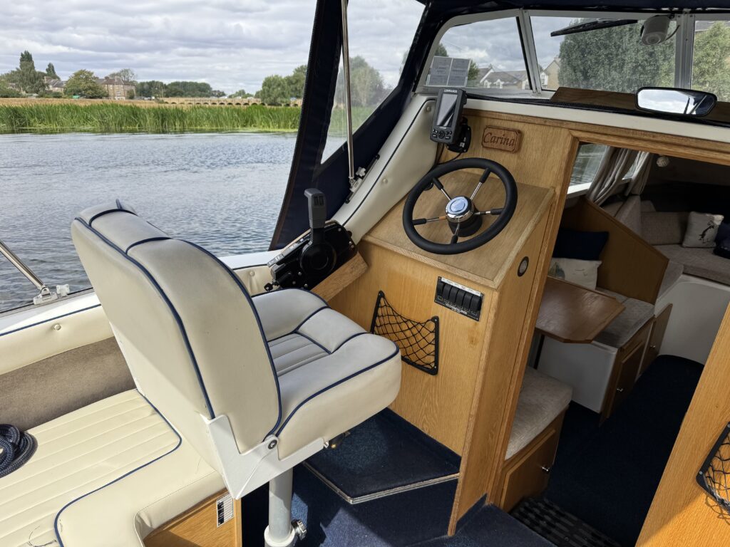 The interior of a boat featuring a white cushioned captain's chair with gray trim, a steering wheel, and a wooden dashboard. The view shows a calm waterway with greenery in the background, visible through large windows.