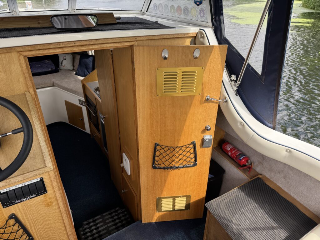 An interior view of a boat showing a wooden storage cabinet with a yellow vent and a grid-patterned pocket. The floor is carpeted, and a steering wheel is visible on the left. Natural light enters from a nearby window, reflecting on the water outside.
