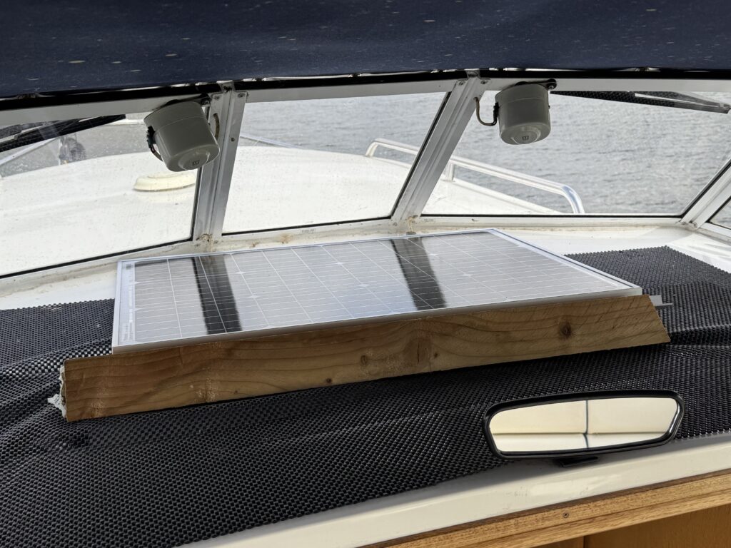 A solar panel mounted on a wooden frame is placed on the dashboard of a boat. The boat's cabin features a dark netted surface and large windows with a view of water in the background.