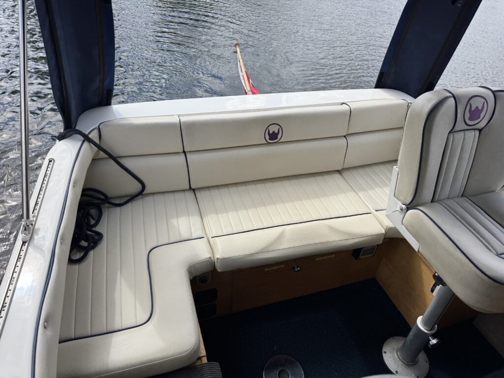 An interior view of a boat featuring a white seating area with padded cushions, including a bench and a captain's chair. The boat's upholstery displays a dark blue trim, and the reflective water is visible in the background.