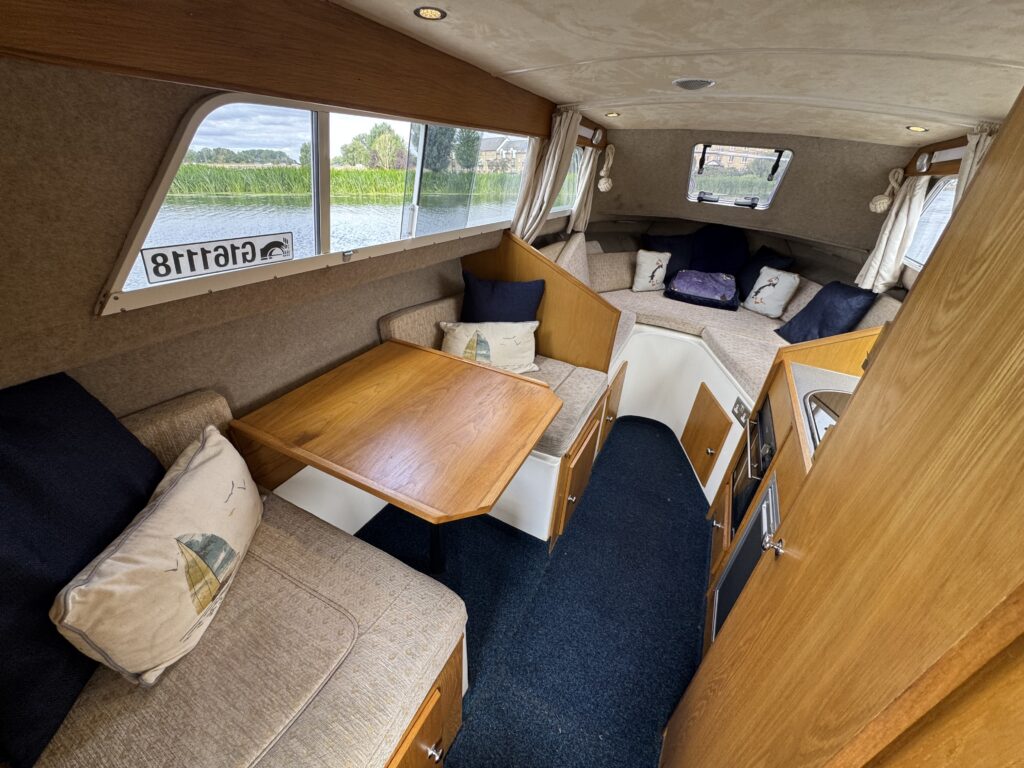 The interior of a small yacht features a wooden table surrounded by cozy seating with cushions. Large windows allow natural light and provide scenic views of the water and greenery outside. The space is designed for comfort while cruising.