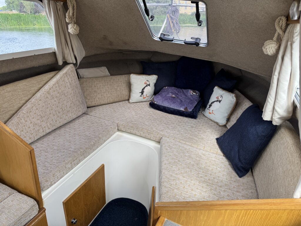 An interior view of a small boat cabin featuring a U-shaped seating area with beige cushions and blue accent pillows. A large window allows natural light to enter, and a small door leads to the cabin's lower section.