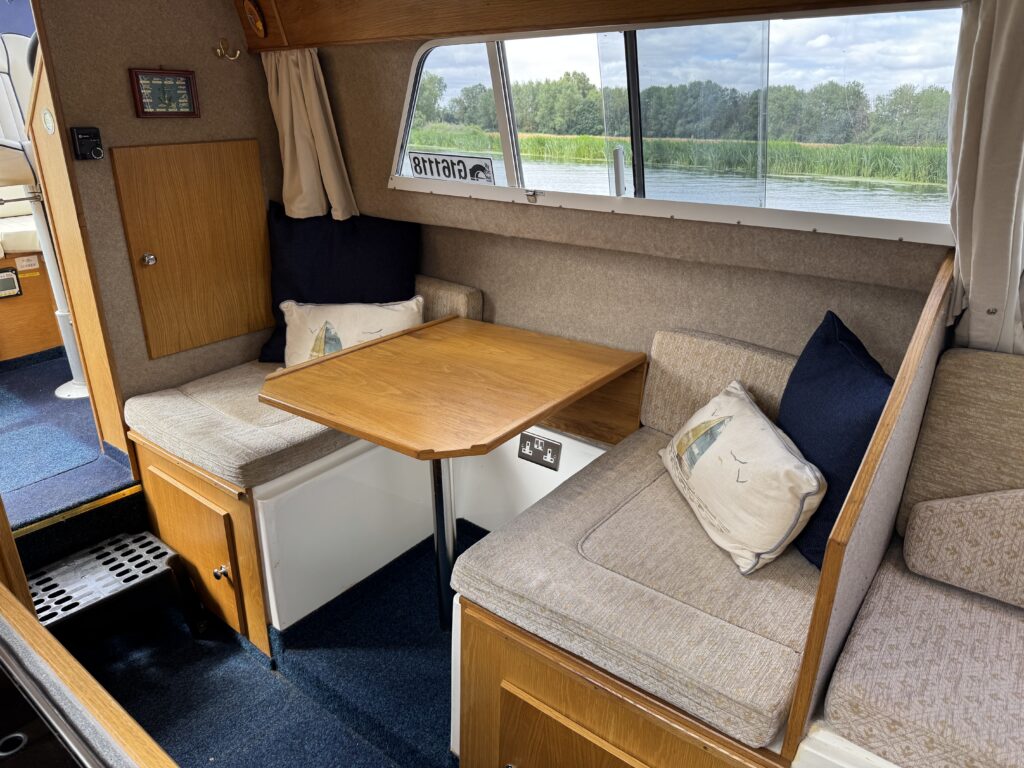 An interior view of a boat cabin featuring a wooden table surrounded by cushioned seating. The cushions are light-colored with decorative pillows. Large windows provide a view of greenery outside, and the floor is carpeted.