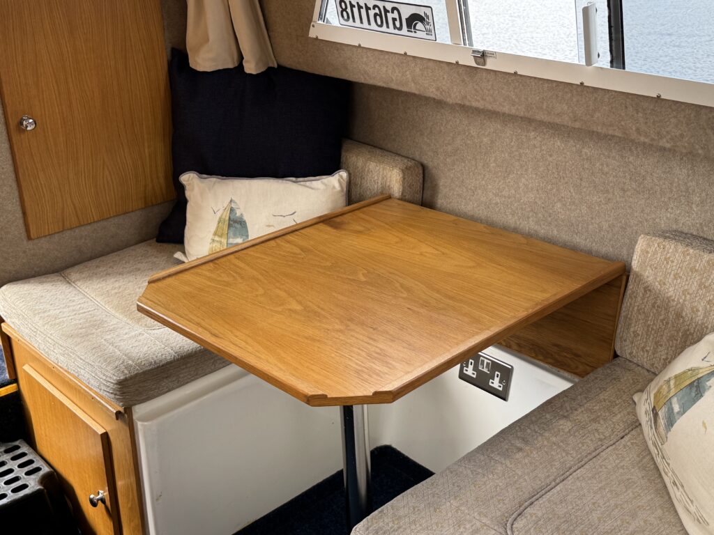 A compact seating area in a boat featuring a wooden table supported by a silver pole. The seating is upholstered in light gray fabric, complemented by a pillow with a sailboat design. The walls are lined with beige carpeting, and natural light comes through a nearby window.
