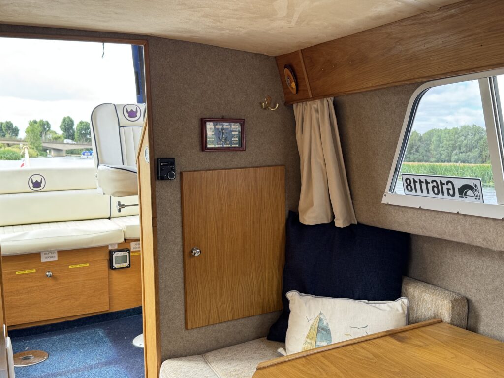 The interior of a boat cabin features a wooden bench seat with a decorative pillow, a small table, and a wall-mounted cabinet. A window reveals the outside view with greenery, and a plush seating area is visible in the background.