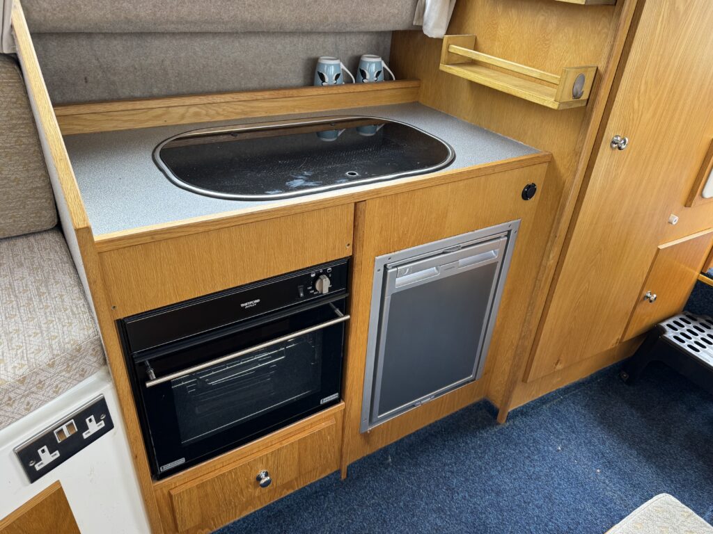 A compact kitchen area featuring a built-in sink with a black countertop, an oven below, and a small refrigerator to the right. The cabinetry is wooden, and there is shelf space above the countertop with two metal cups.