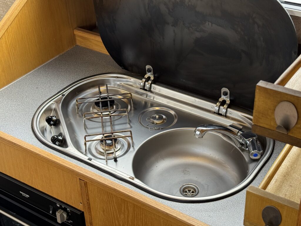 A stainless steel kitchen sink with a built-in two-burner gas stove is displayed under a countertop. The stove features black knobs for ignition, and a small faucet with a blue lever is positioned beside the sink.