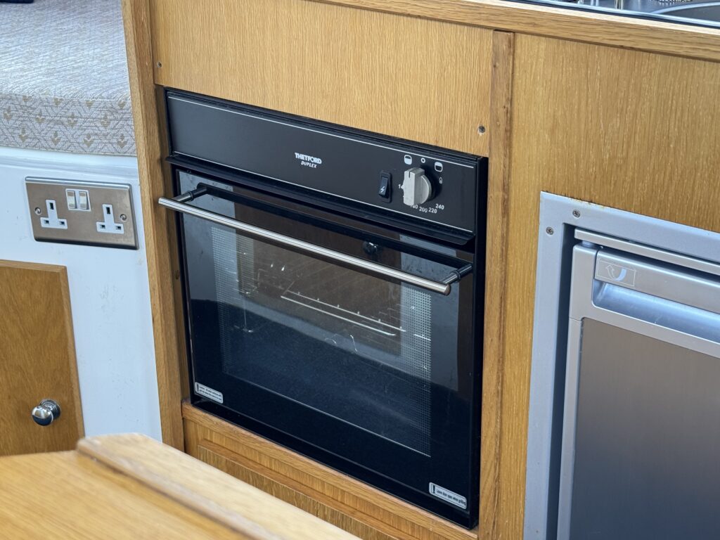 A built-in black oven with a glass door located within wooden cabinetry. The oven features a control knob and a handle, positioned next to an electrical outlet and a silver-colored appliance, likely a refrigerator, on the right.