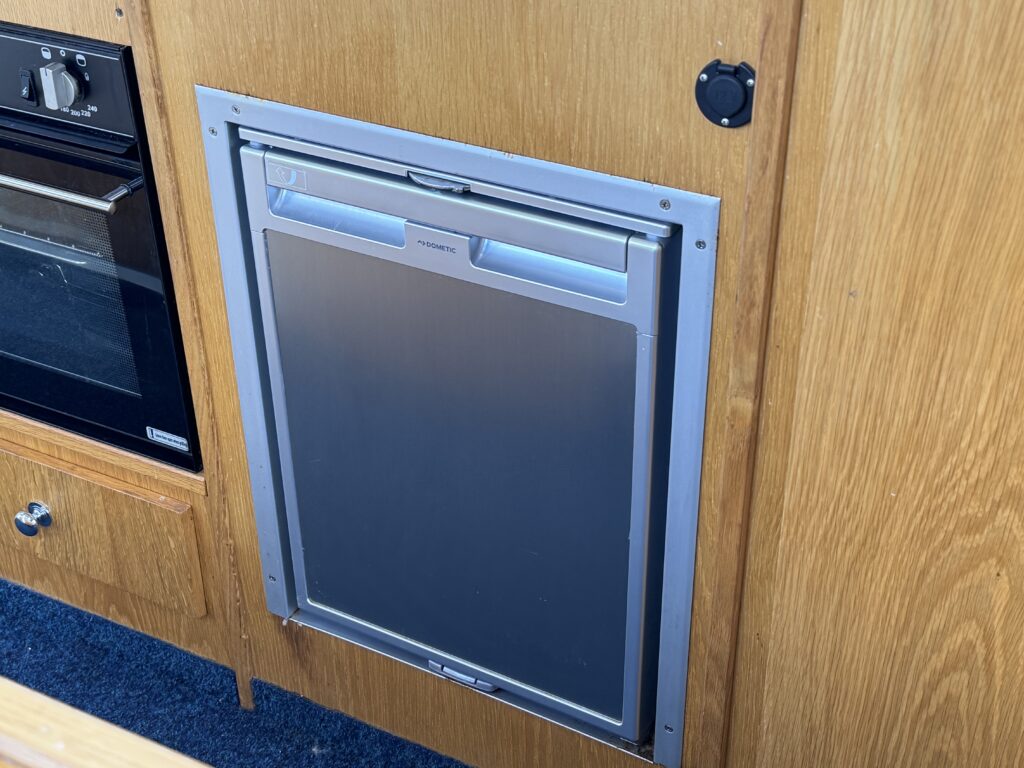 A stainless steel refrigerator door installed in wooden cabinetry, featuring a sleek handle and a control panel at the top. The surrounding area shows light-colored wood and dark blue carpet flooring.