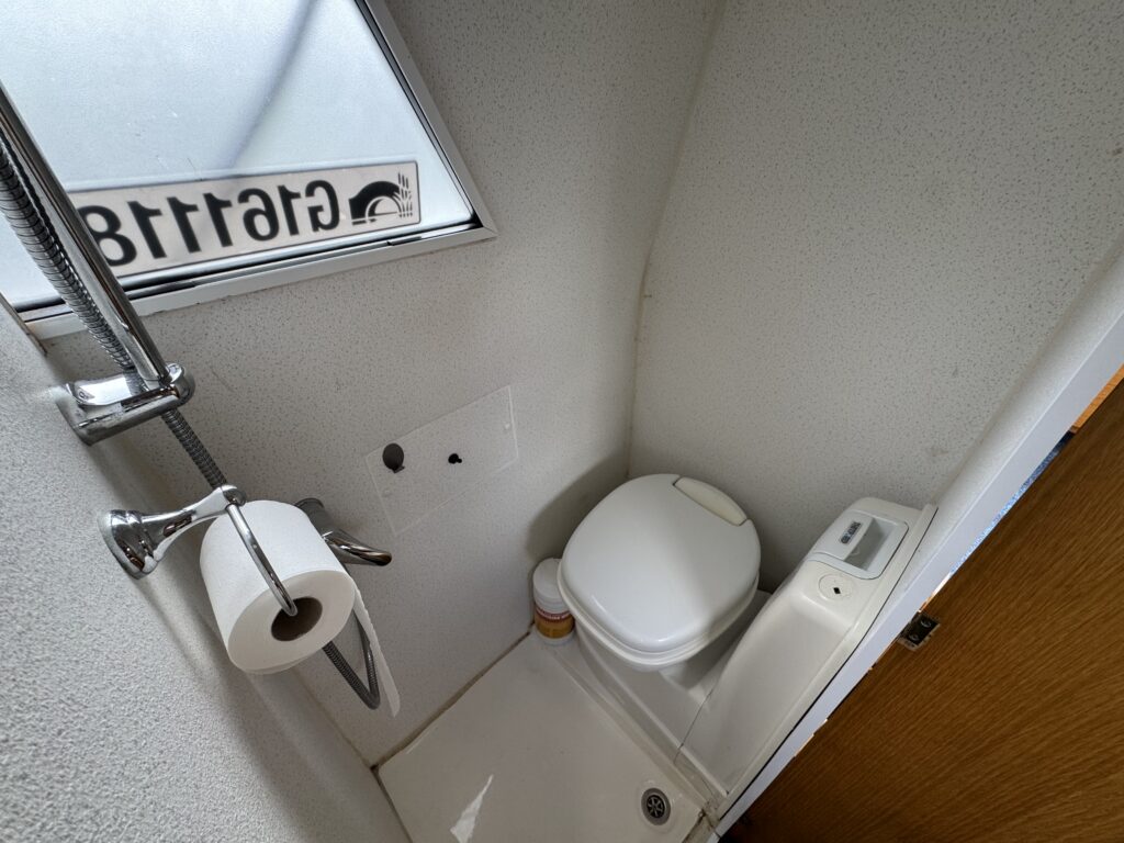 A compact bathroom space featuring a white toilet, a small shower area, a toilet paper holder, and a mirror. A window above the toilet provides natural light with a view of the exterior, while walls are finished with a light texture.