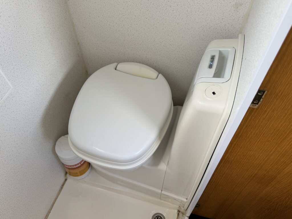 A white toilet positioned in a compact bathroom corner with a visible control panel on the side. The surrounding walls are light-colored, and there is a closed wooden door in the background.