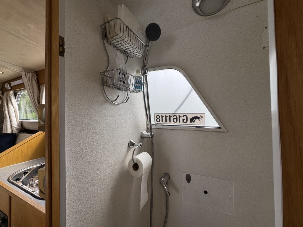 A shower stall in a compact space, featuring a shower head, a small shelf with toiletries, and a roll of toilet paper on a holder. The wall has a small window with a sign indicating "Gratitude" visible through it.