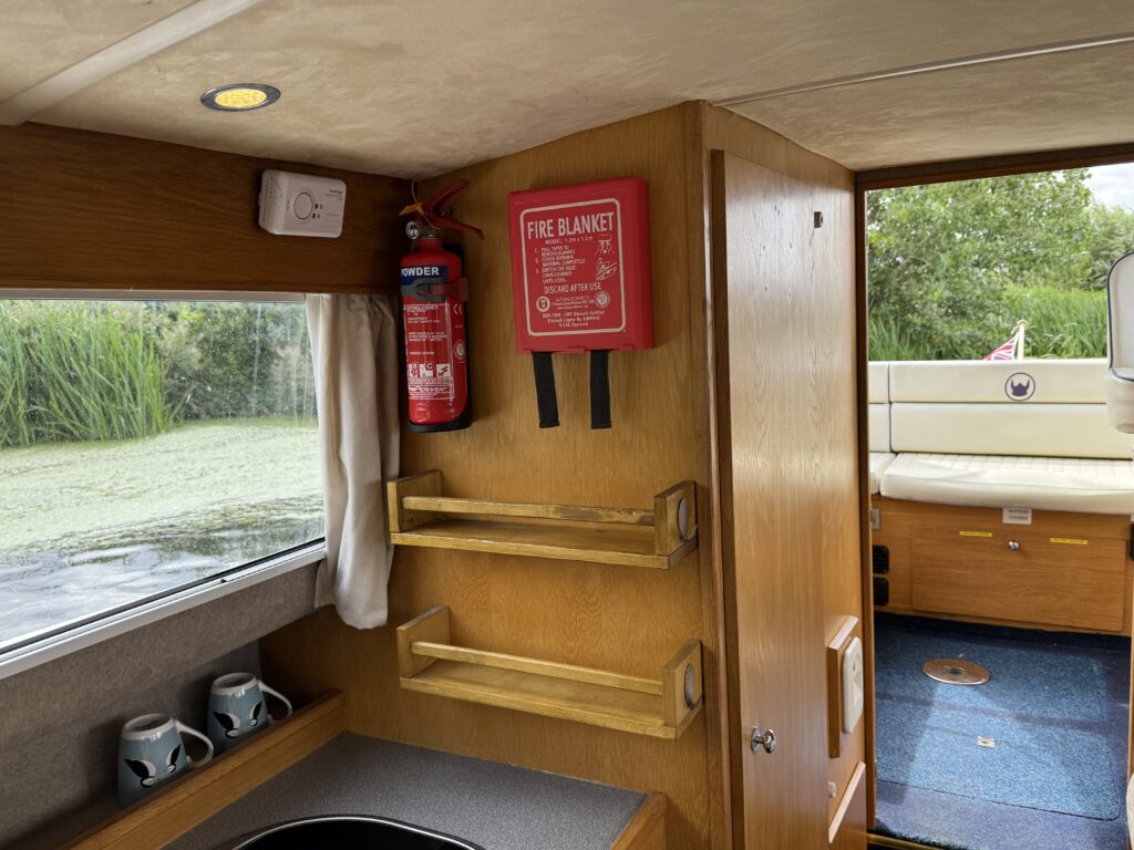 An interior view of a boat showing a wooden cabinet with a fire blanket mounted on the wall and a fire extinguisher beside it. Below, there are two wooden shelves, and a gray countertop is visible in front of a large window with greenery outside.