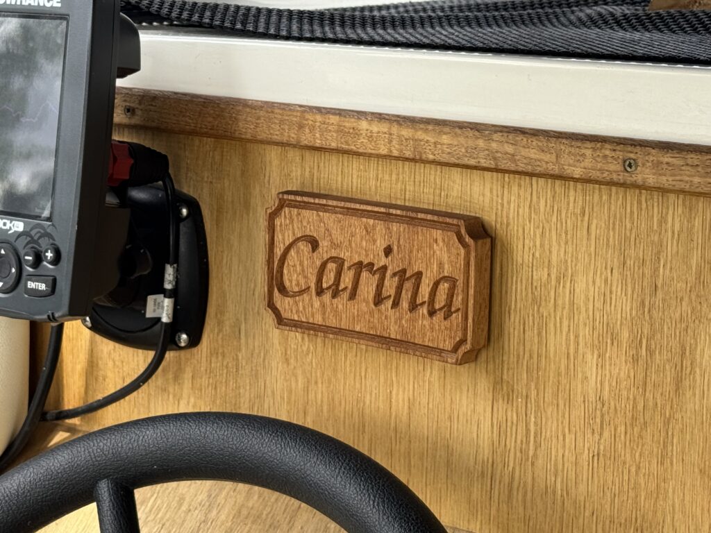A wooden plaque with the name "Carina" carved in a cursive font, mounted on a wooden surface. To the left, a fish finder is attached to the wall, and a black steering wheel is visible in the foreground.