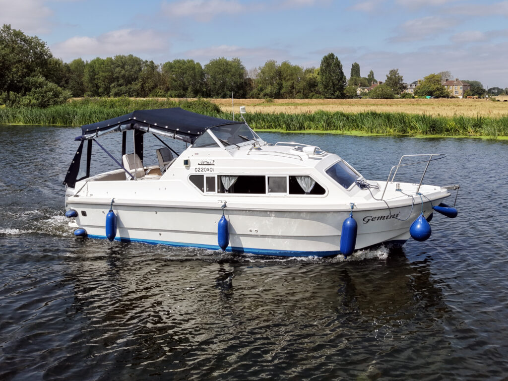 A white cruising boat named 'Germind' with a blue stripe is navigating on a calm waterway. It features a blue canopy, and two blue fenders are attached to the sides. Lush greenery and a building can be seen in the background.