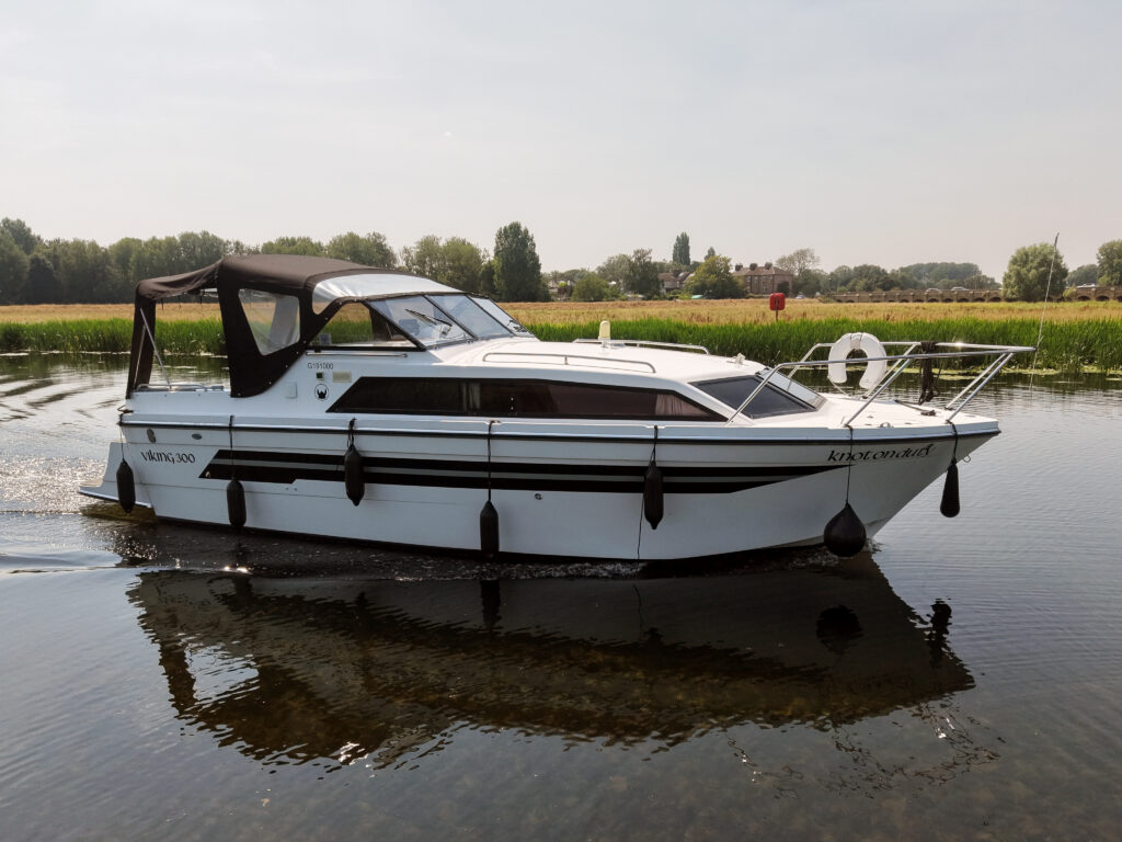 A white motorboat with a black canopy and stripe moves through calm water. Surrounding the boat are green trees and open fields under a clear sky.