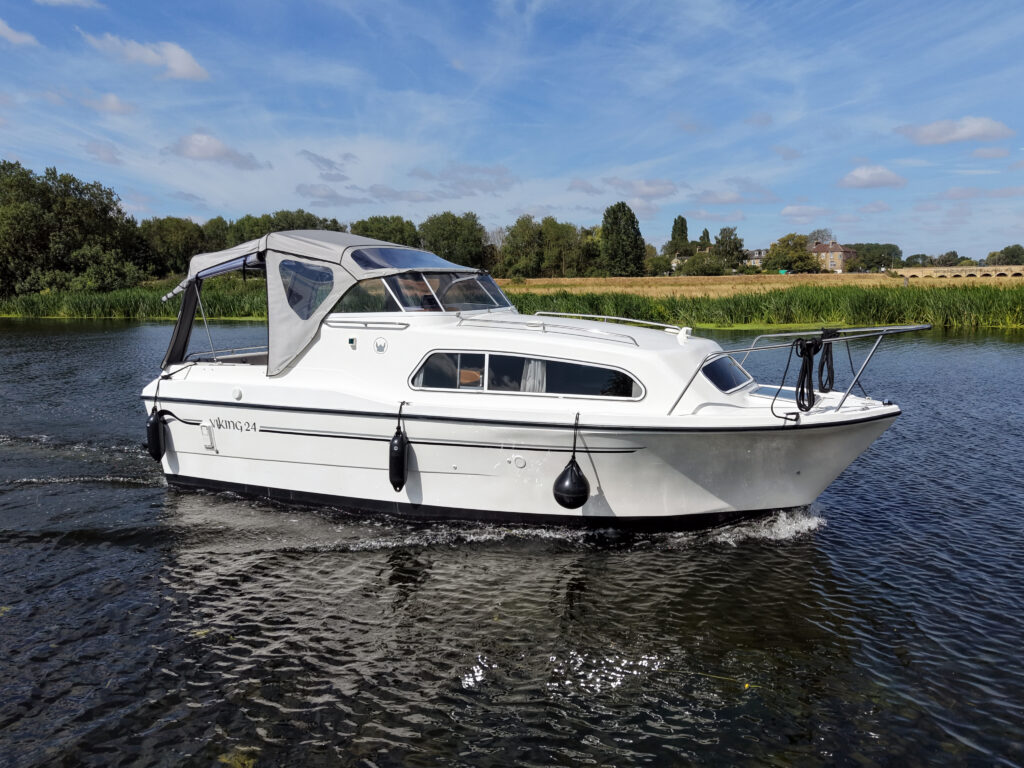 A white motorboat with a cabin is cruising on a calm river. The background features lush greenery and a clear blue sky with scattered clouds.