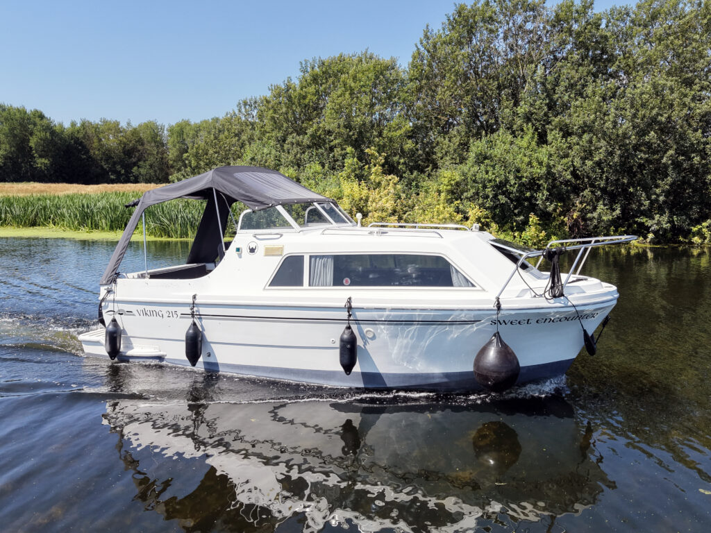 A white motorboat with a gray canopy is cruising on calm water. The background features green trees and shrubs along the shoreline.