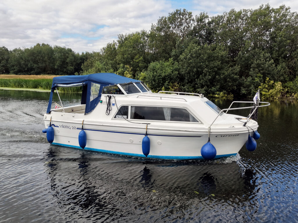 A white motorboat with a blue canopy and fenders, cruising on calm water. In the background, lush greenery and trees line the shore under a partly cloudy sky.