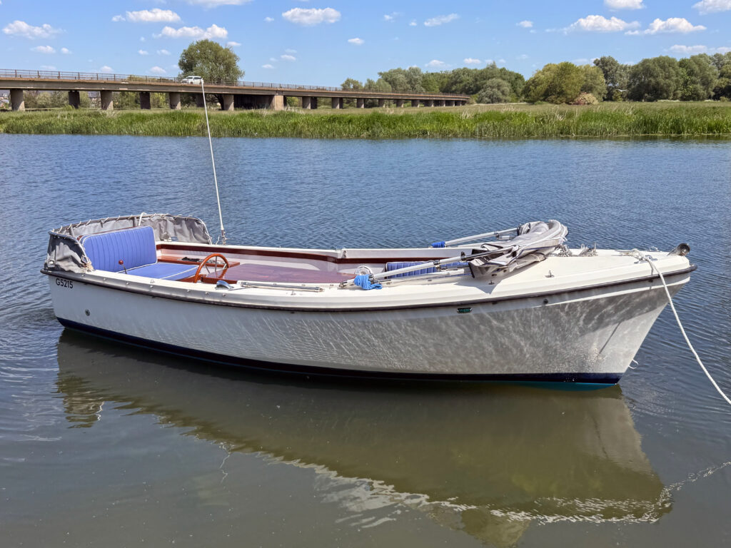 A white motorboat with a beige canopy is moored on a calm river. The boat has blue seating and a wooden steering wheel, with a bridge visible in the background and green vegetation along the riverbank under a clear blue sky.