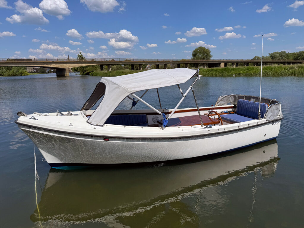 A white motorboat with a blue canopy is moored on calm water. The boat features blue cushioned seating and a wooden steering wheel. In the background, a bridge spans over the river, with green vegetation lining the water's edge.