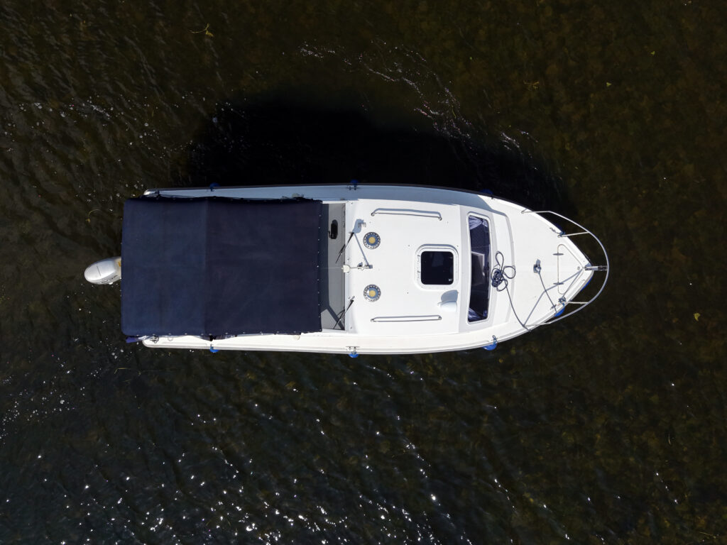 An overhead view of a white boat with a dark blue canopy on a greenish-brown body of water. The boat features several round metal fittings and a small window on the cabin.