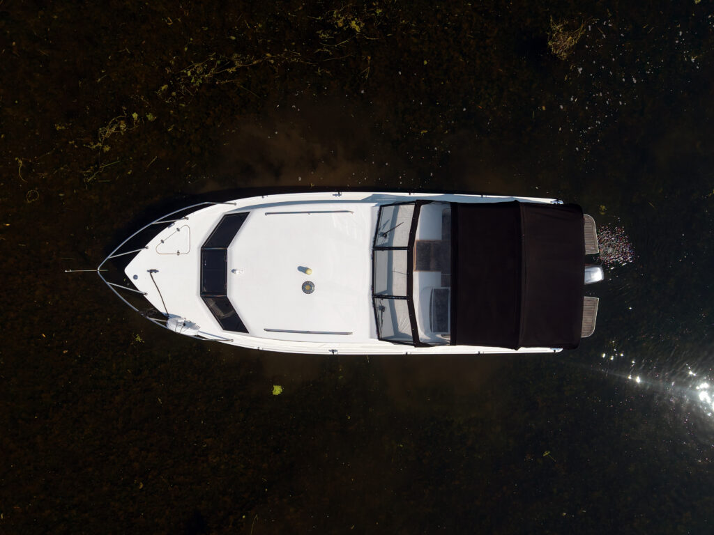 An aerial view of a white motorboat with a black canopy floating on dark water. The boat features a streamlined design and a small deck area at the back, with patches of greenery visible beneath the water's surface.