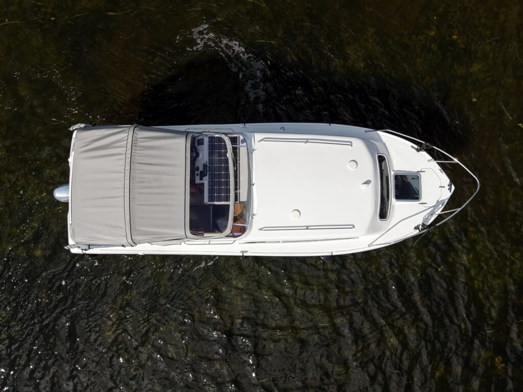 An overhead view of a white motorboat with a canvas cover parked on calm water. The boat features a solar panel on the roof and is surrounded by a natural green environment.