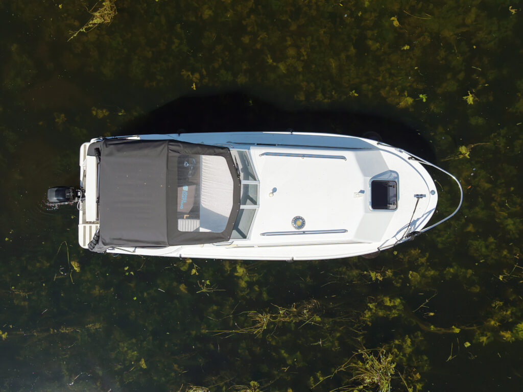An aerial view of a white motorboat with a black canvas cover on top, floating on clear water. Below the surface, green aquatic plants are visible, contrasting with the boat's exterior.