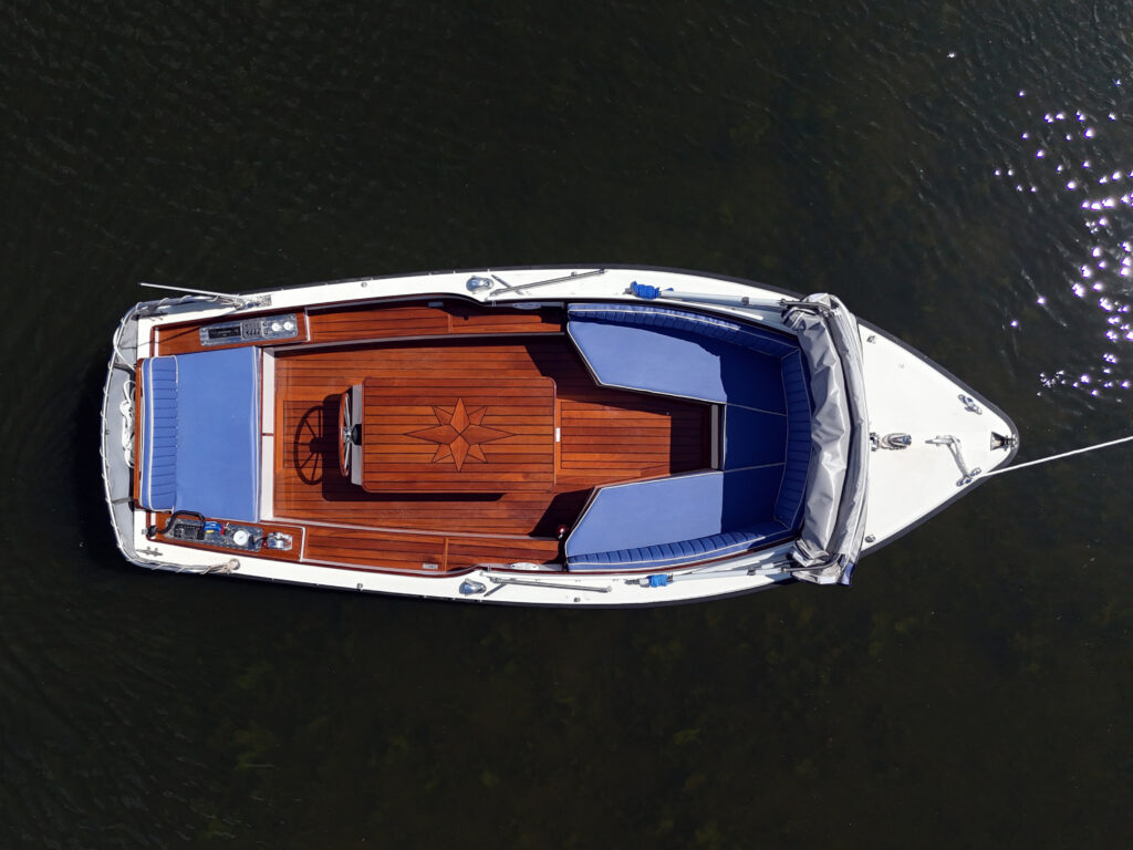 An aerial view of a boat with a white hull and a sleek design, featuring a wooden deck and blue seating. The boat is anchored on calm water, reflecting sunlight, with a steering wheel visible at the center.