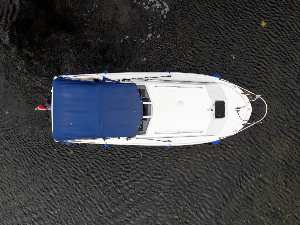 An overhead view of a white motorboat with a blue canopy, resting on dark water. The boat has a small windshield and is secured with blue straps, featuring a flag at the back.