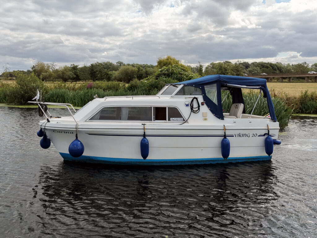A white motorboat named 'Viking 20' is moored in a calm waterway. The boat features a blue canopy and fenders, with greenery and trees visible in the background under a cloudy sky.