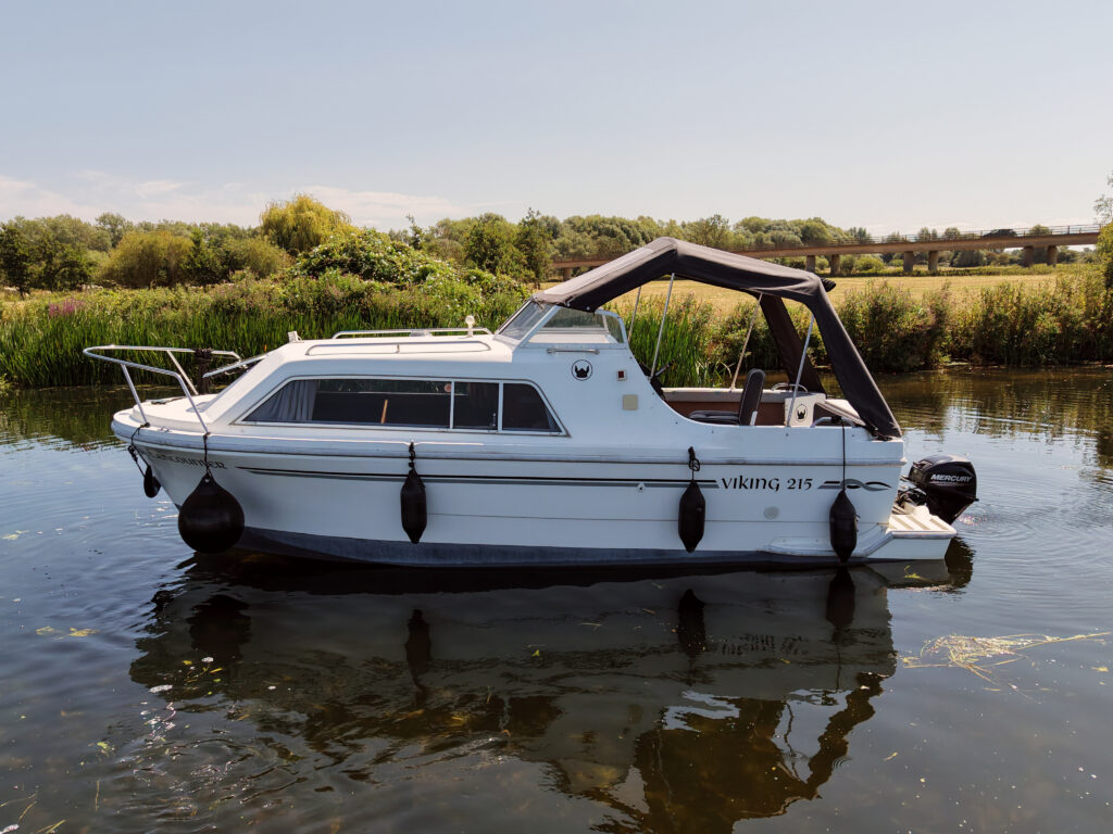 A white Viking 215 boat is moored on calm water, surrounded by green foliage. The boat has a black canopy and is equipped with fenders on both sides, with a grassy bank visible in the background.