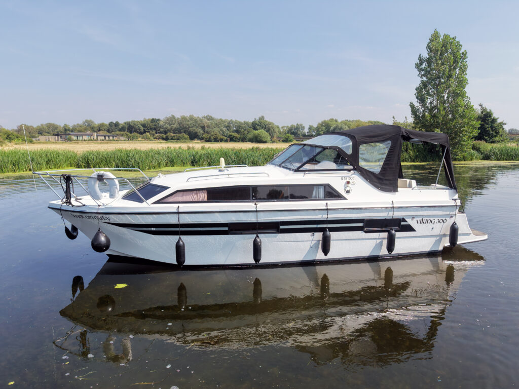 A white motorboat with a black canopy is anchored on calm water, with reflections visible on the surface. Lush green vegetation and trees line the background under a clear blue sky.