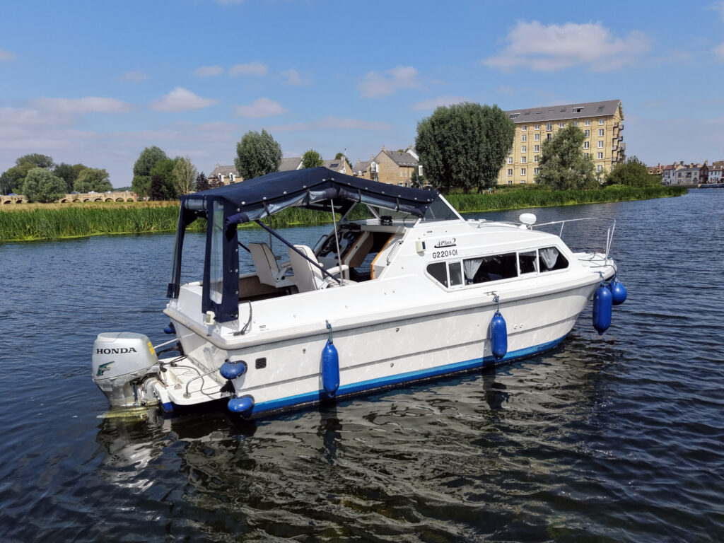 A white motorboat with a blue stripe and a canopy is anchored in a river. The boat has two blue fenders and an outboard motor labeled "Honda." The background features trees and a building along the riverbank under a partly cloudy sky.