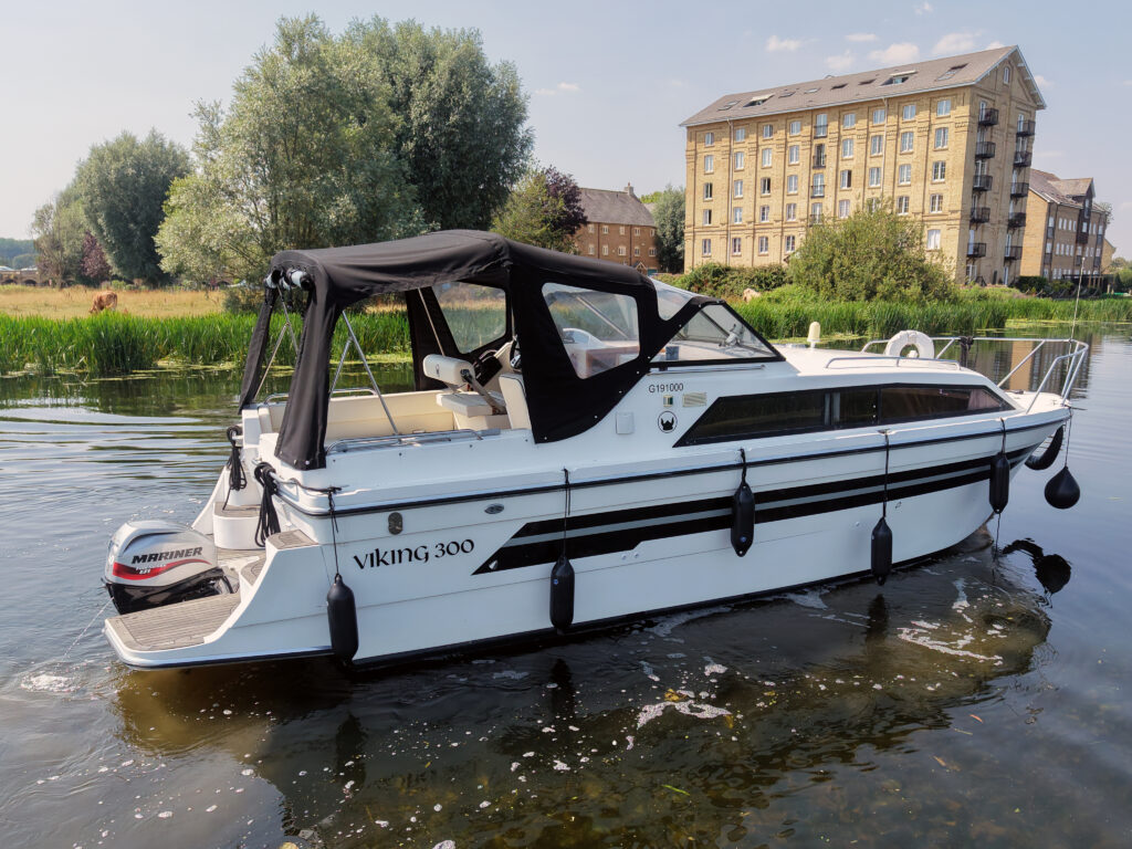 A white Viking 300 boat with a black canopy is docked on calm water. In the background, a tall building and trees provide a natural landscape, reflecting the warm sunlight.