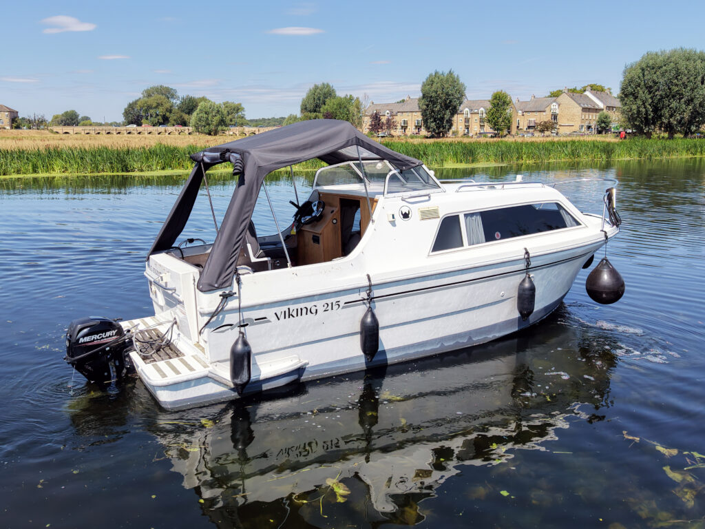 A white Viking 215 motorboat with a gray canopy is moored on calm water. Surrounding the boat are green trees and fields, with blue sky and clouds visible in the background.