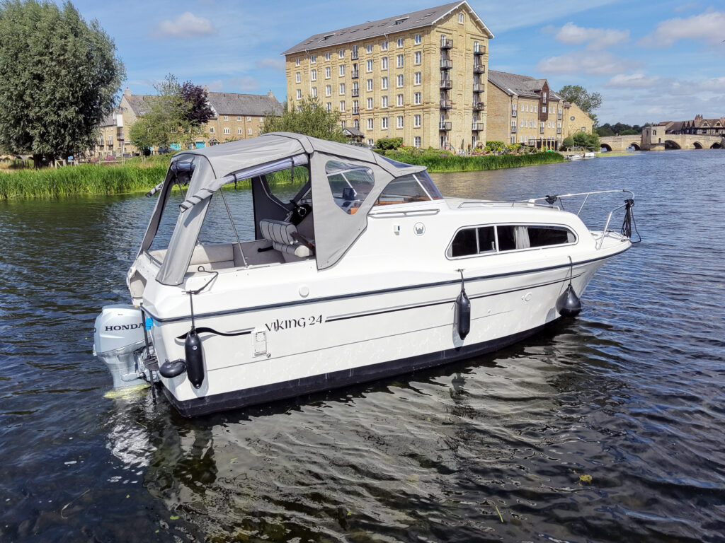 A white Viking 24 motorboat with a gray cockpit cover is docked on calm waters. In the background, a brick building with large windows is surrounded by trees and greenery, under a blue sky with scattered clouds.