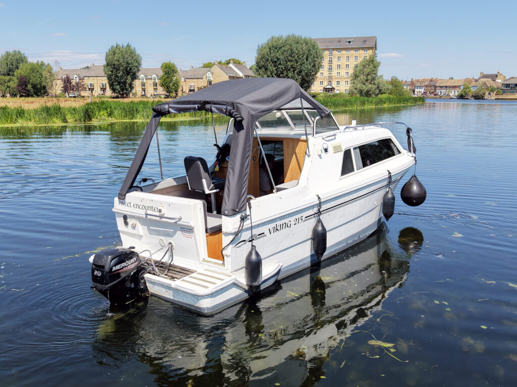 A white motorboat named 'Viking 217' is floating on calm water, with its canopy open. The boat has an outboard motor attached at the rear and is surrounded by greenery and a low brick building in the background.