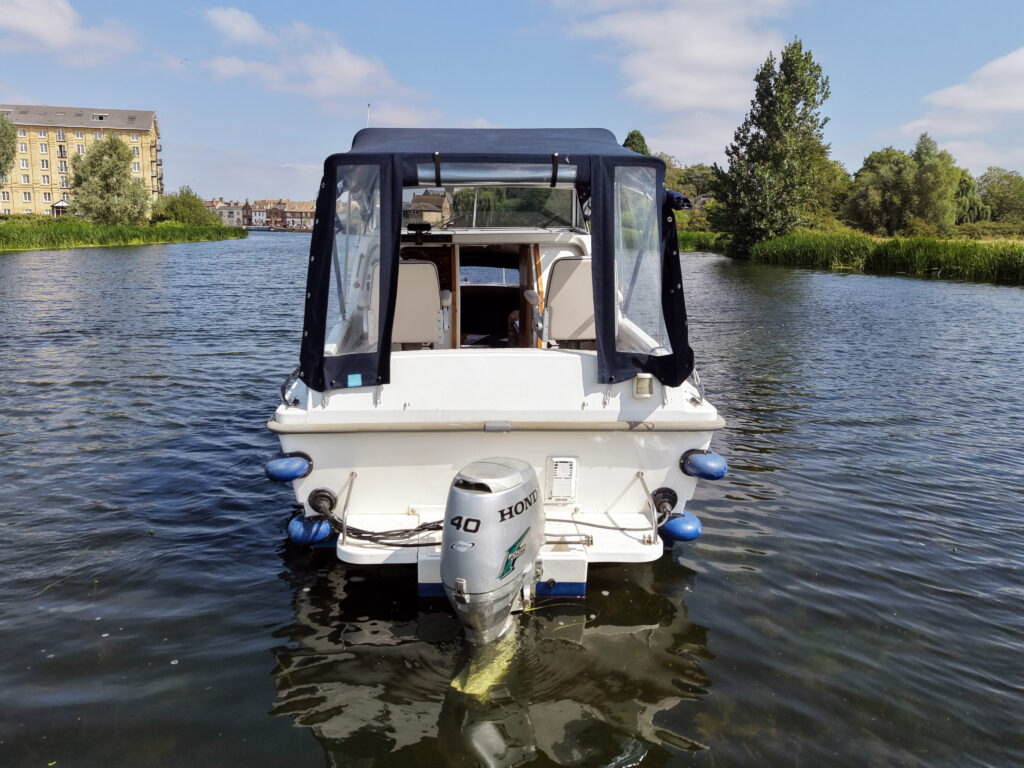 A white motorboat with a blue canopy is positioned on calm water. The boat features a Honda outboard motor, and the background includes a riverside building and green trees under a partly cloudy sky.