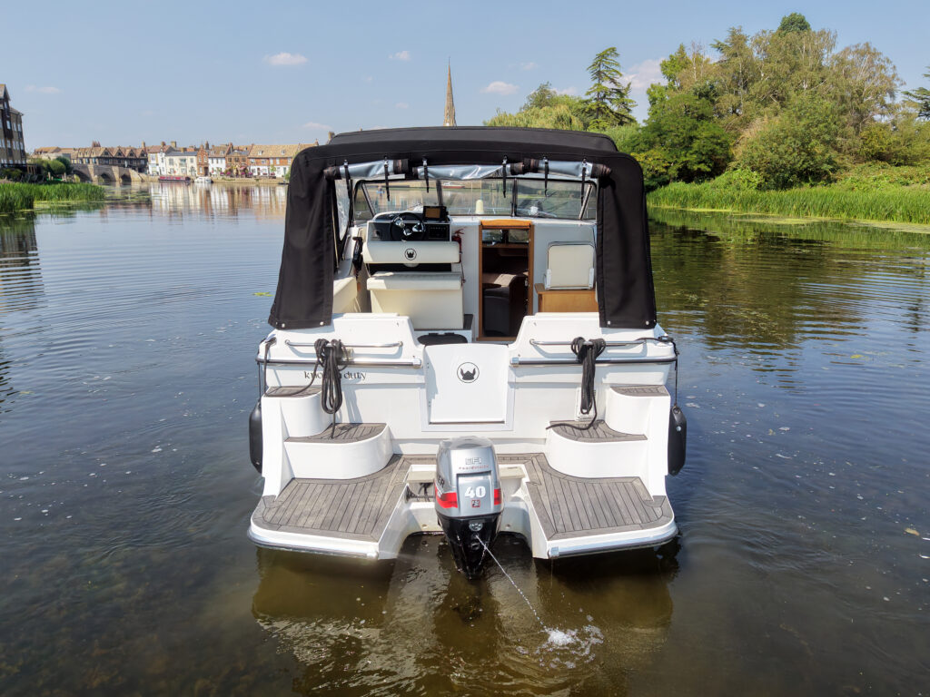 A white motorboat with a black canopy is anchored in a calm river. The rear view shows the boat's engine and a spacious seating area, with trees and a distant church steeple visible along the riverbank in the background.