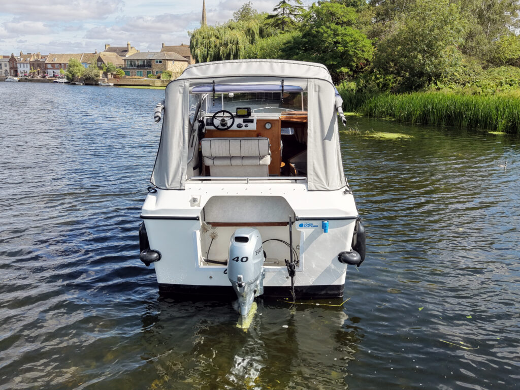 A white motorboat with a gray canopy, seen from the rear, is moored in calm water. The boat shows a control panel at the helm and has a 40 HP outboard motor, with landscaped greenery and houses visible on the shore in the background.