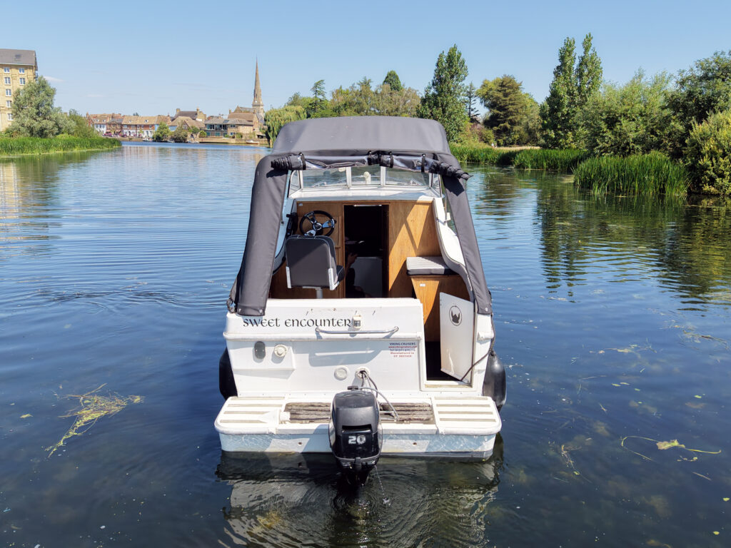 A small white boat with a black canopy is anchored on calm water. The boat features a rear motor and wooden interior elements. In the background, there are trees and a visible steeple from a church, under a clear blue sky.