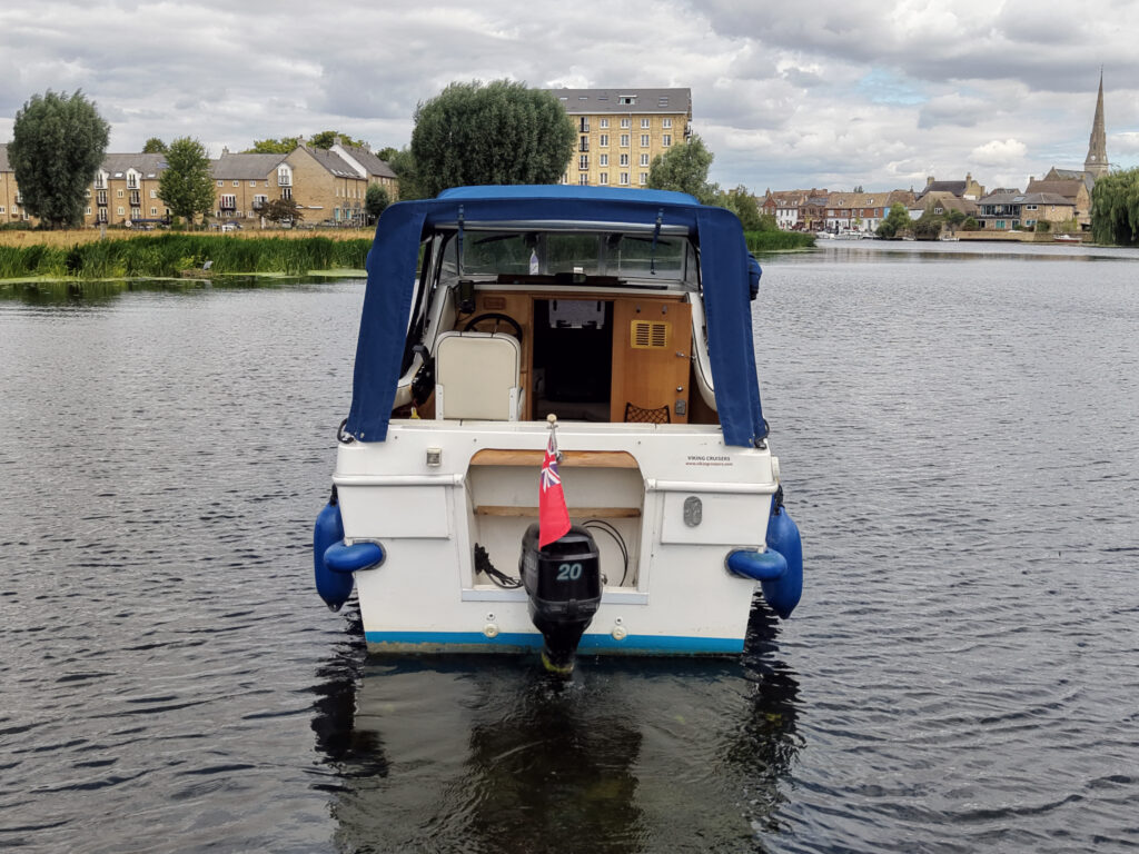 A white and blue motorboat with a blue canopy is anchored in a calm body of water. The boat's rear features a black motor and a small pink flag, while buildings and trees are visible in the background under a cloudy sky.
