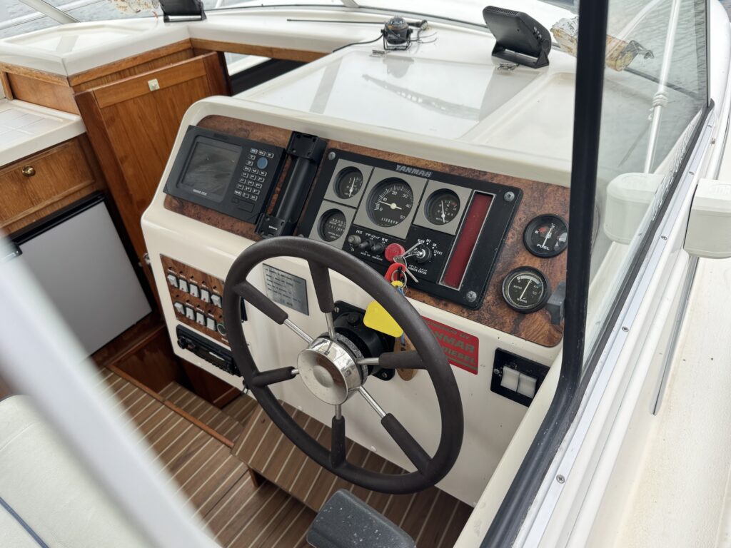A boat helm station with a steering wheel, analog gauges, and a radio panel, set beside wood cabinetry and a striped teak-style floor.
