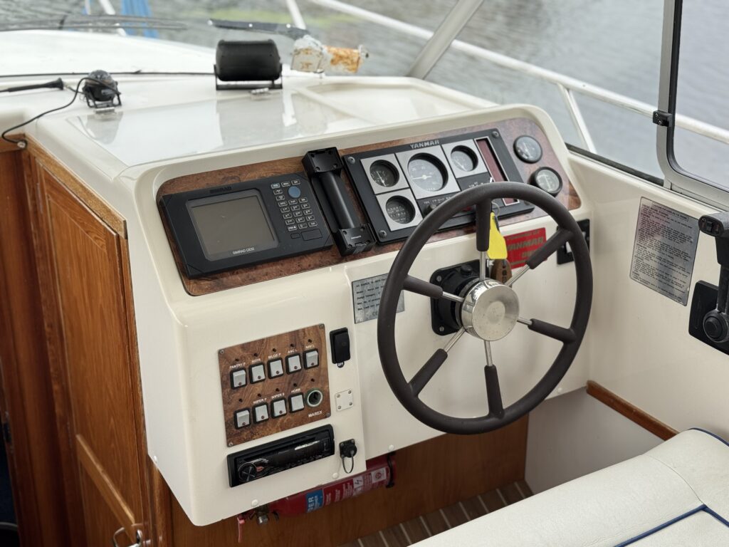 A boat helm with a steering wheel, gauges, and control switches, set against a white dashboard and wood trim.