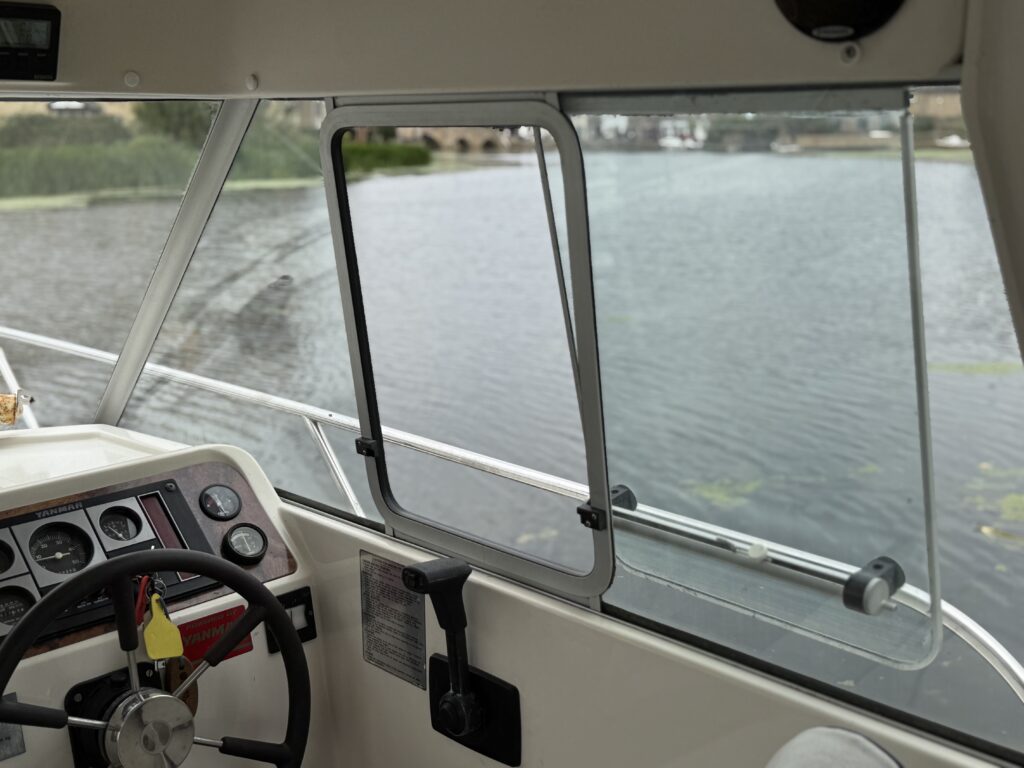 The helm of a Marex 280 Holiday boat with a steering wheel and control panel beside large windows overlooking calm water.