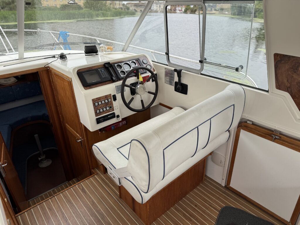 The helm of a Marex 280 Holiday boat with a steering wheel, instrument panel, and white cushioned bench seat beside large windows overlooking the water.