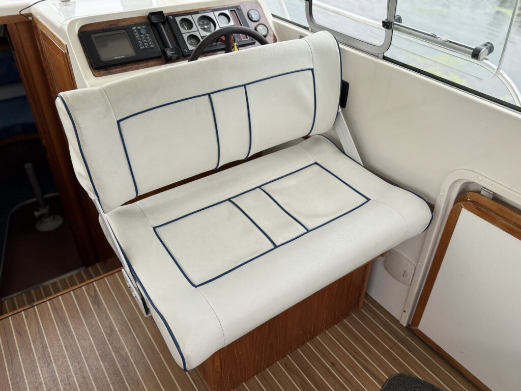 A cream-upholstered boat bench seat with navy piping sits beside the helm console, with dashboard controls, steering wheel, and wood-trimmed interior visible.